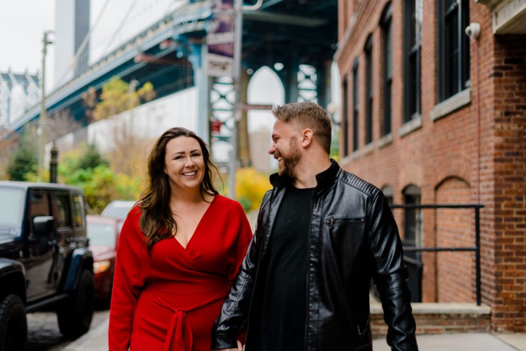 woman in a red dress and a man in a black leather jacket walking in DUMBO together. The man is looking over at the woman smiling. 