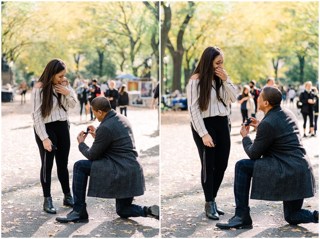 man kneeled down on one knee proposing to his girlfriend in central park. She has her hand covering her face.