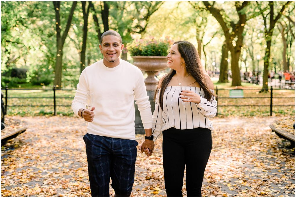 a couple walking in central park holding hands. The woman is looking over at the man with a smile on her face. 