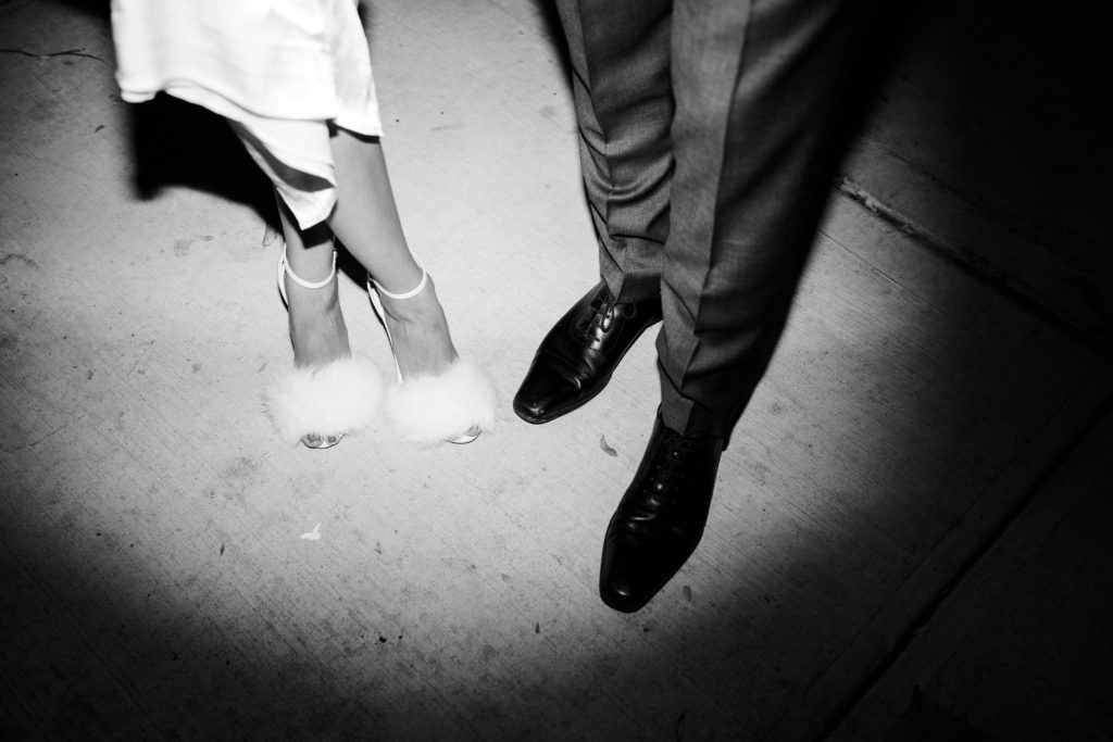 a pair of white heels and feet standing next to a pair of black shoes. They are lit up in the shape of a spotlight.