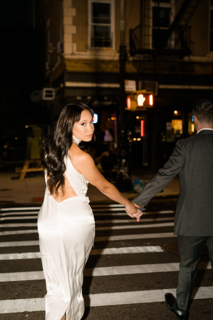 couple walking on a crosswalk while holding hands. Woman in a white dress is looking over her shoulder