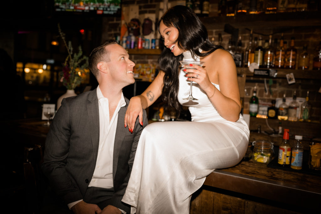 woman in a white dress sitting on the counter of a bar leaning on a man in a grey suit. They are looking and smiling at each other.