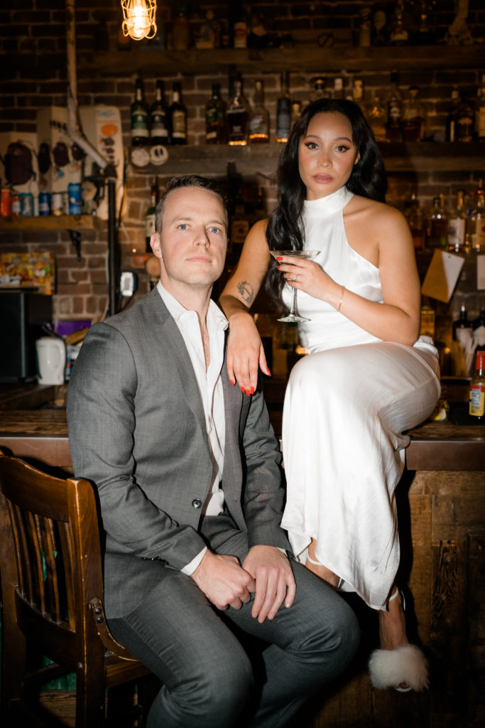 woman in a white dress sitting on top of a bar next to a man in a grey suit who is sitting on a bar chair
