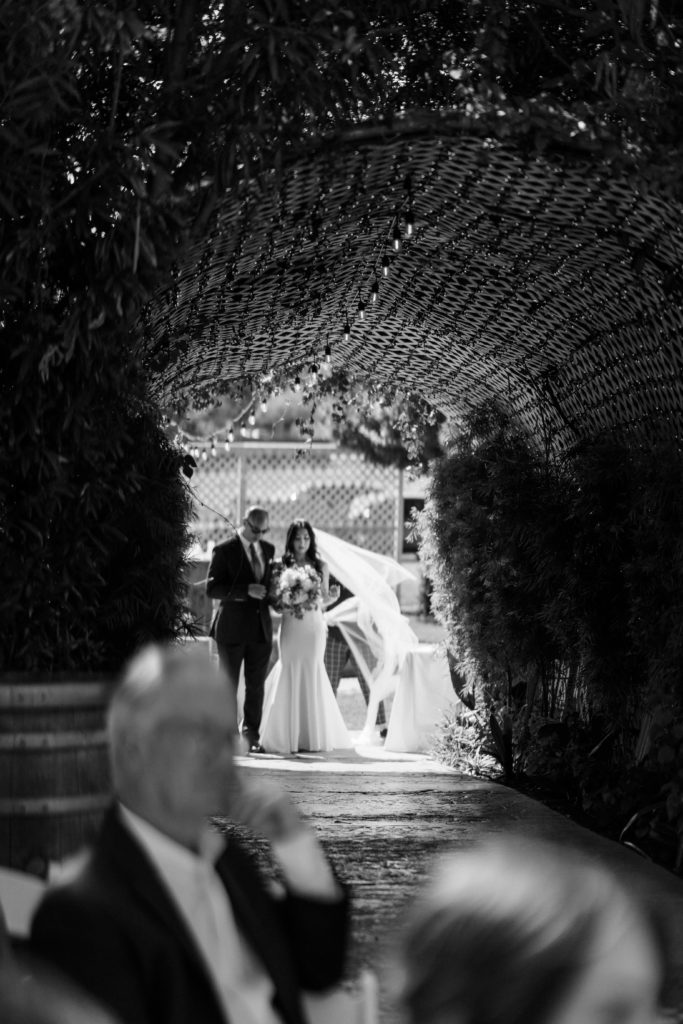 A father walking his daughter down the aisle under a tunnel. The brides veil is blowing in the wind behind her