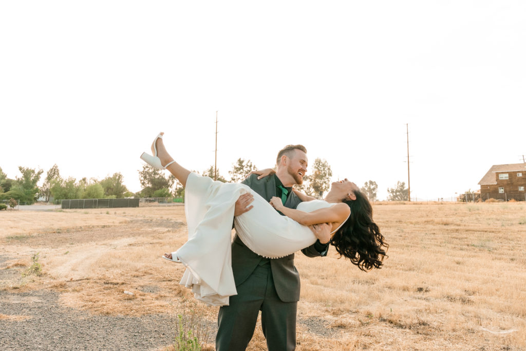 a groom cradle carrying a bride in a field. They are laughing together.