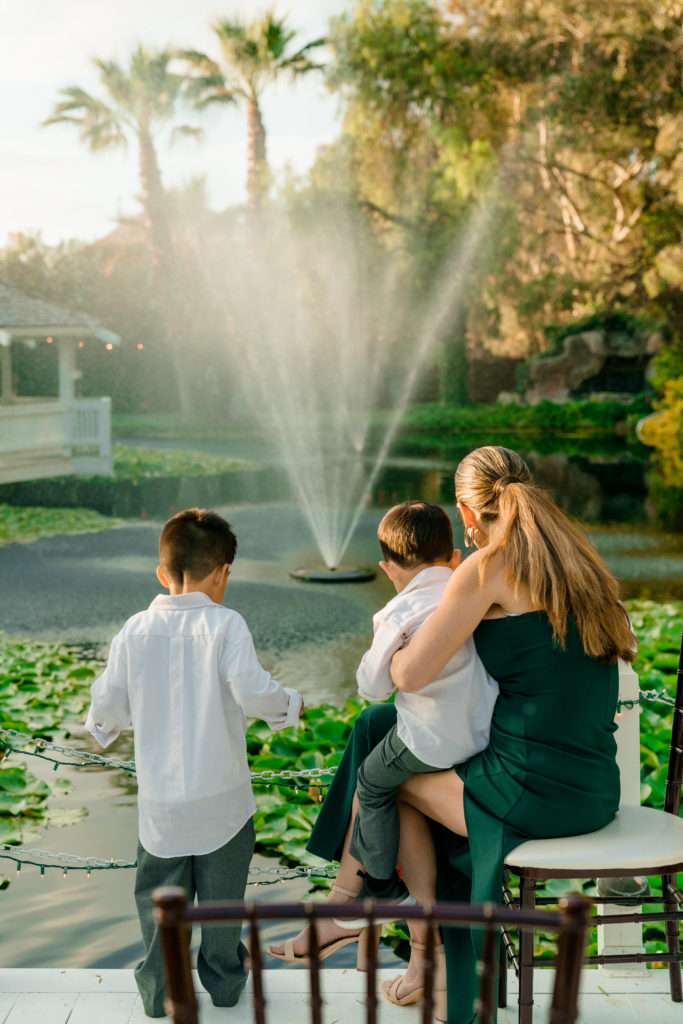 a woman holding a little boy on her lap while another boy stands next to them. They are facing away looking at a pond with a fountain