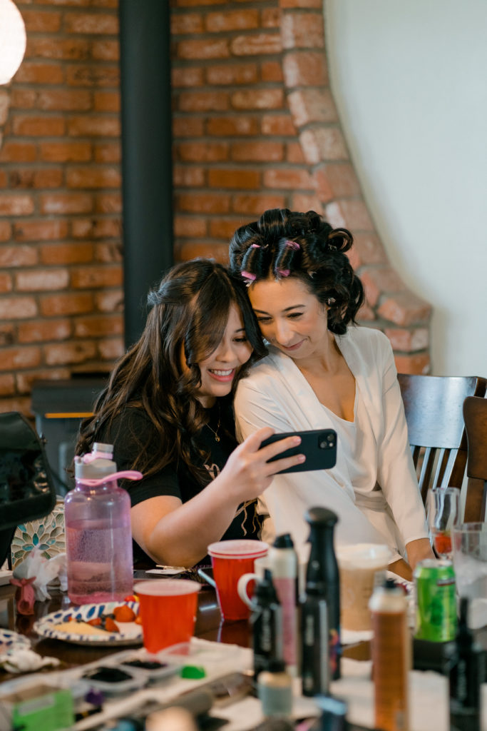 a woman with curlers in her hair sitting on another womans lap while they take a selfie on a cell phone