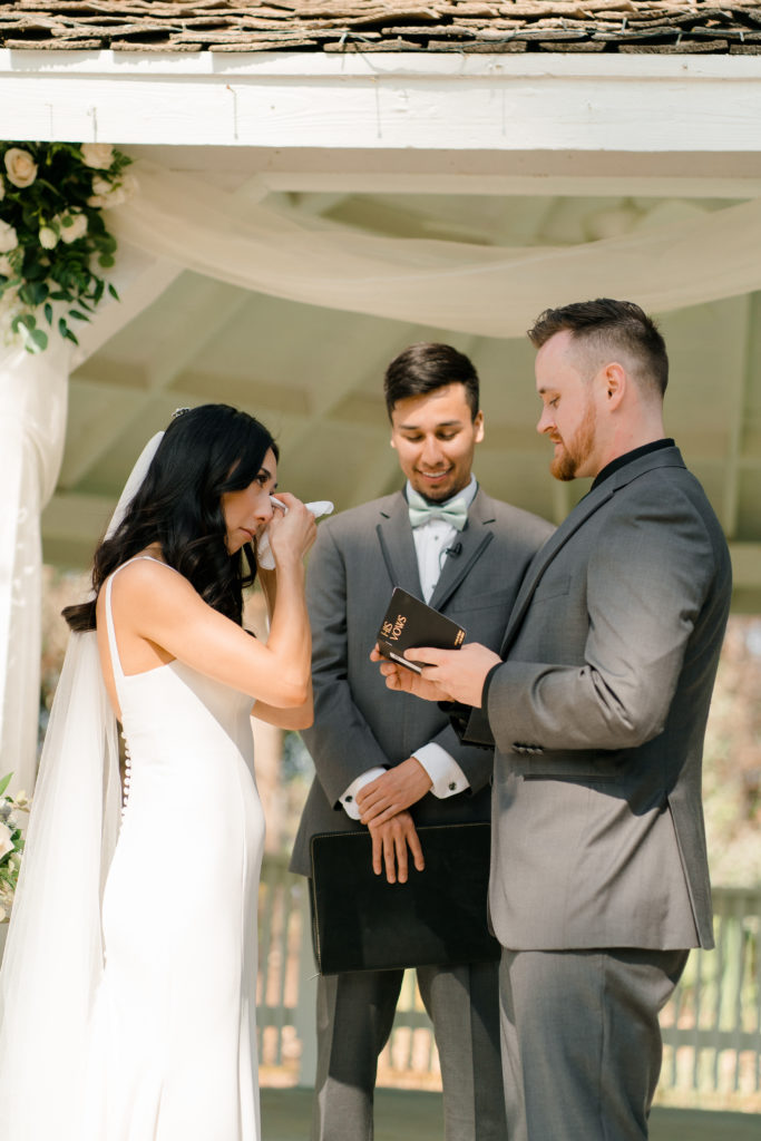 A bride wiping a tear away with a tissue facing the groom. The groom is looking down at a little book while he is talking