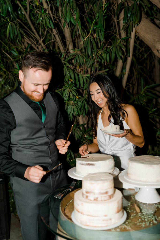 a bride and groom laughing together while they stand behind 3 wedding cakes