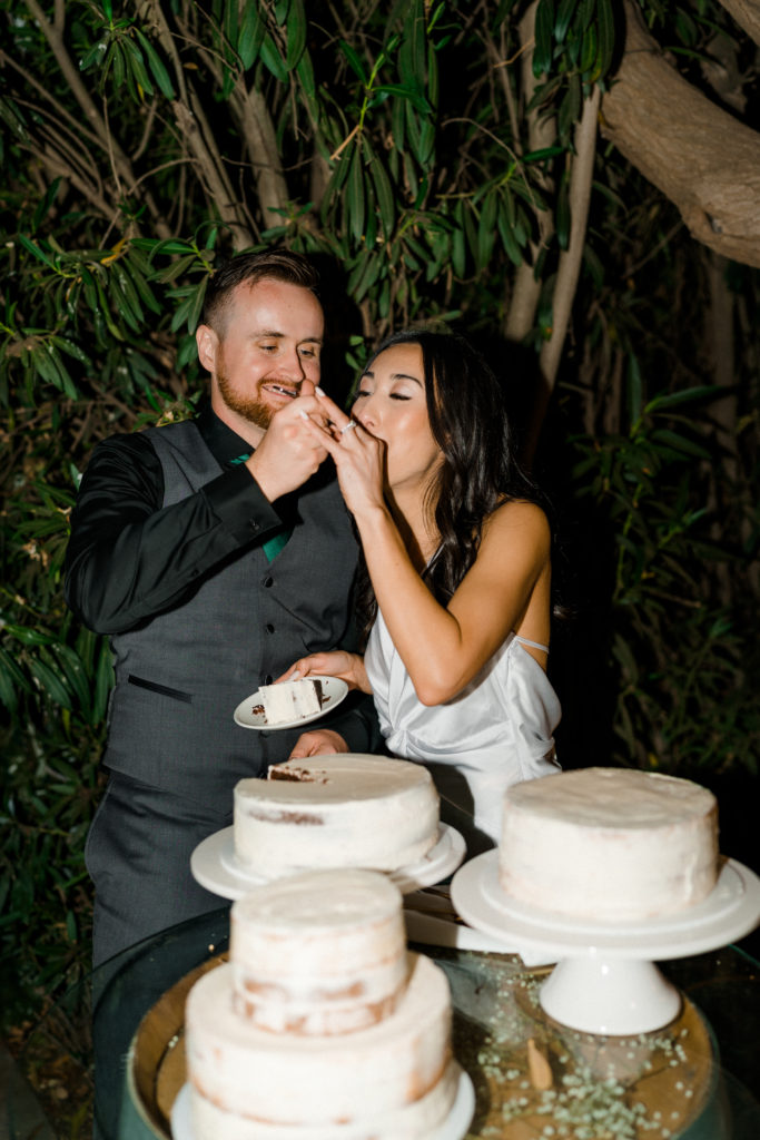 a bride and groom standing behind a table of three wedding cakes. The groom is feeding the bride cake.