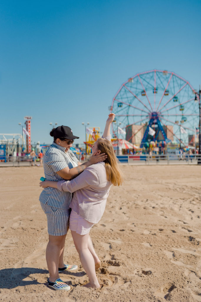 a couple celebrating in front of the coney island ferris wheel. One person is holding the other persons face. 