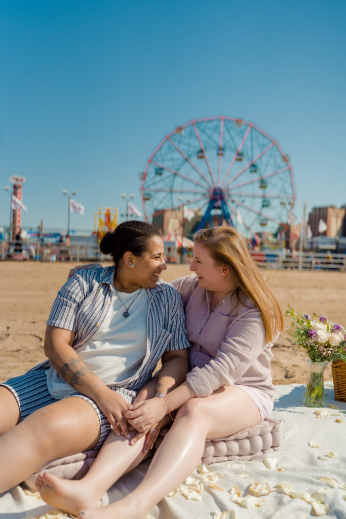 two women sitting on a beach blanket while they are laughing and looking at each other. They are sitting in front of a ferris wheel