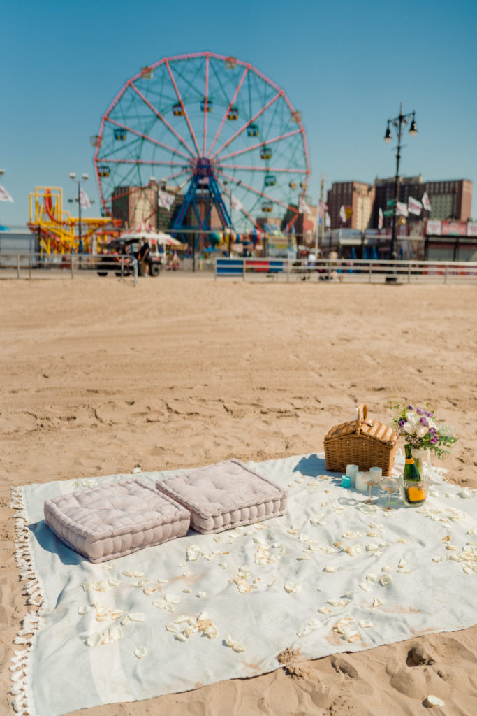 a beach blanket with pillows and a picnic basket set up on the beach in front of a ferris wheel