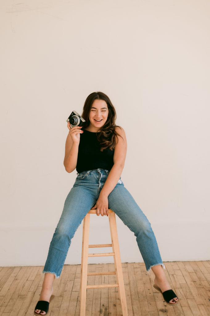 woman sitting on a wooden bar stool while holding a camera in one hand and her other hand is on the stool 