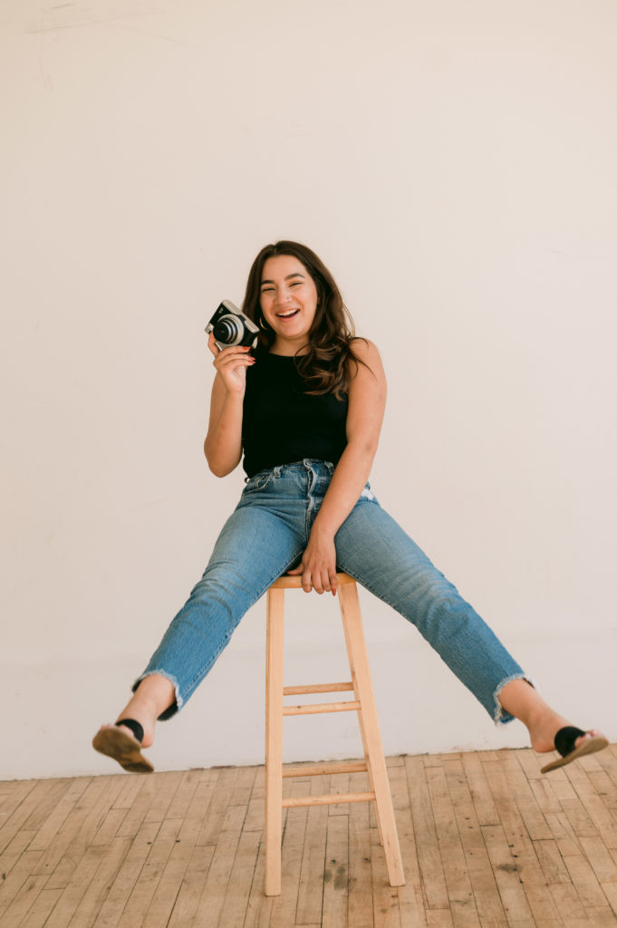 woman sitting on a wooden bar stool holding a camera in one hand and laughing