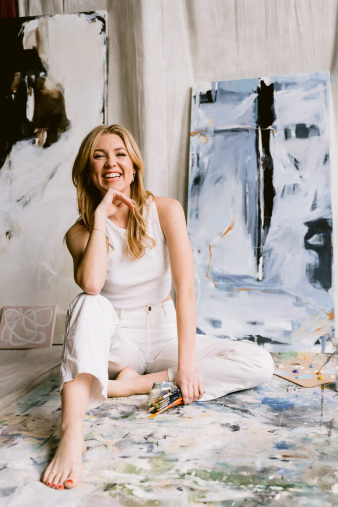 Woman sitting on the ground in an art studio smiling with a painted canvas behind her. 