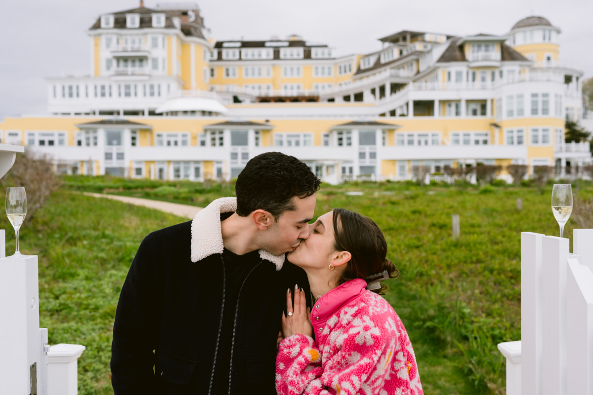 A surprise proposal photograph of a couple kissing in front of a large oceanside building.