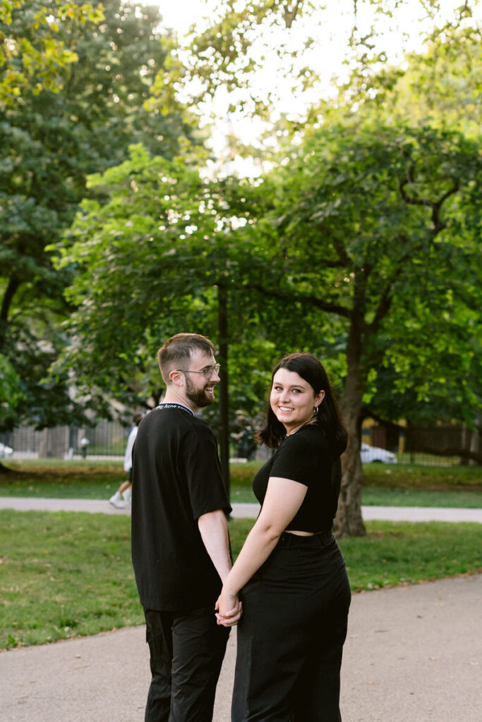A couple walking in a park holding hands as they look back over their shoulders. 