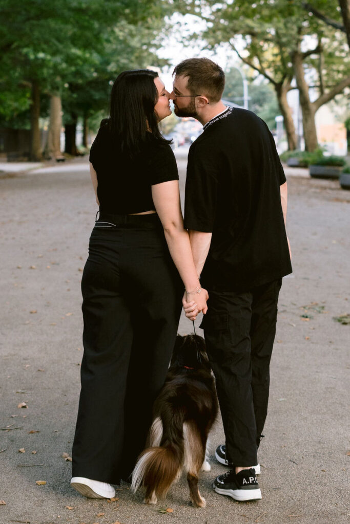 A couple walking in a wooded park holding hands. They are leaning in for a kiss with their dog walking in between their legs below them. 