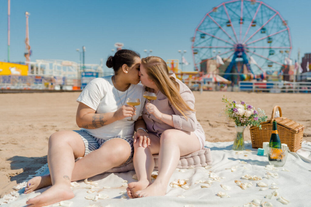 A couple sitting on a blanket on a beach kissing with champagne glasses in their hands. Behind them is Coney Island. 