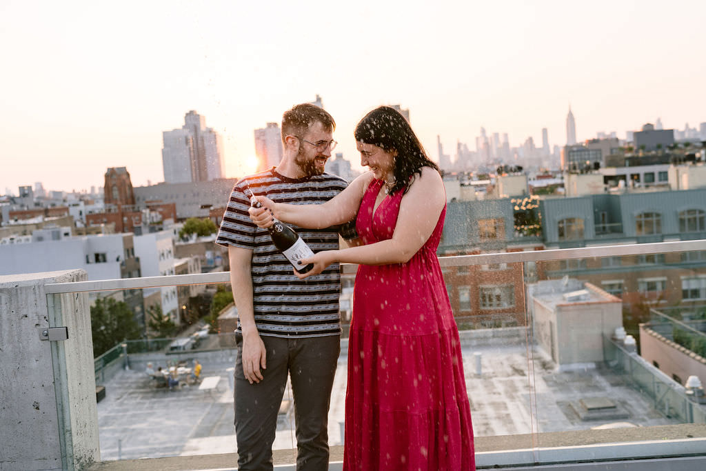 A Brooklyn engagement photo of a couple standing on a rooftop together popping a bottle of champagne.