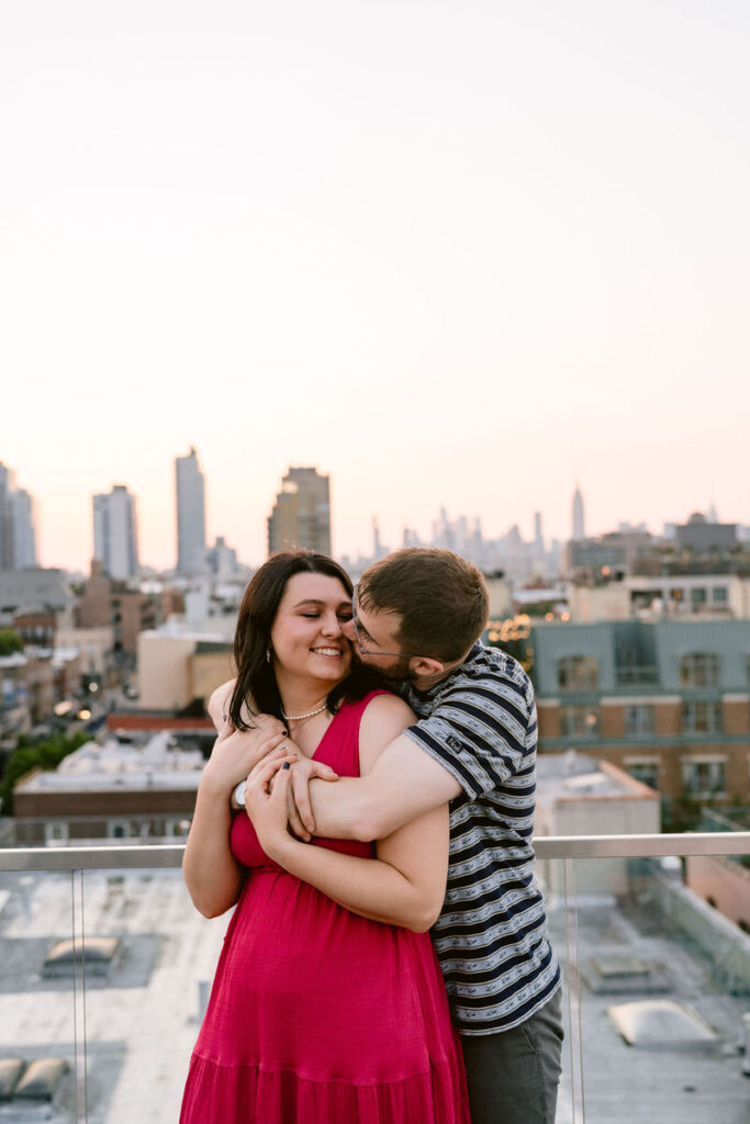A man with his arms around a woman from behind as he kisses her. In the background is the Brooklyn skyline. 