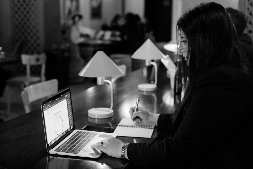 A black and white photo of a woman sitting at a desk working on a laptop and writing in a notebook. 
