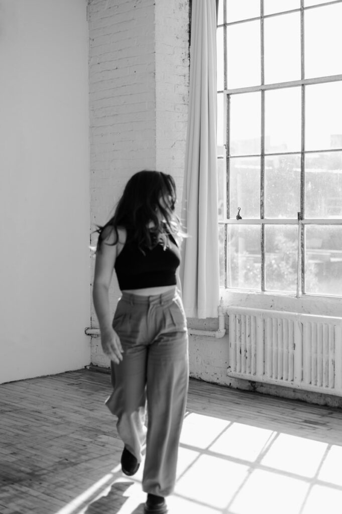 Back and white photo of a woman walking in a loft while looking back out a window. 
