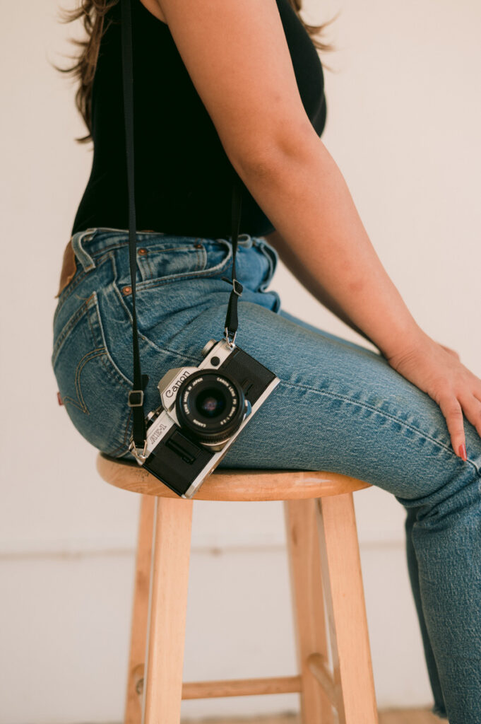 A close up view of a camera hanging off of a person's shoulder as they sit on a stool. 
