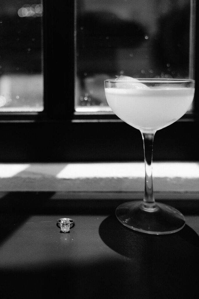 A drink in a tall glass sitting on a windowsill with an engagement ring next to it. 
