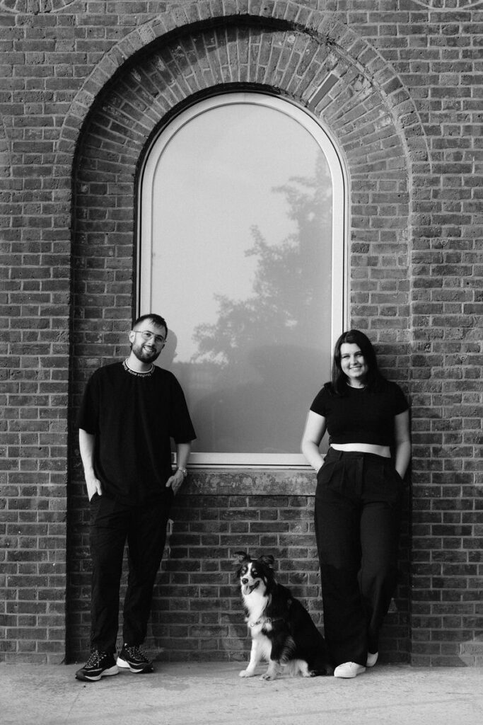 Black and white photo of a couple standing in front of a brick wall with an arched window and their dog is sitting at their feet. 