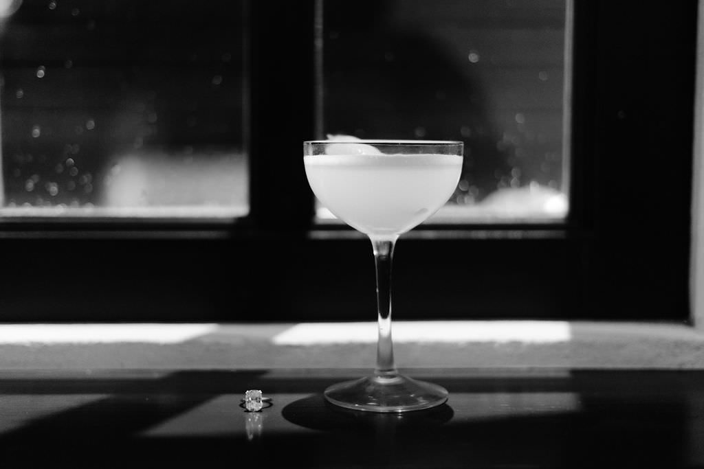 Black and white engagement photo of a drink glass on a windowsill with an engagement ring sitting next to it.