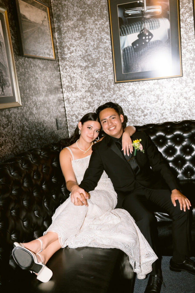 A bride and groom lounging on a black couch together. 

