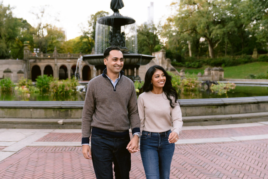 A couple holding hands and walking in a plaza with a fountain in the middle. 