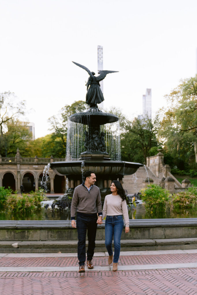 A couple hold hands and smiling as they walk in front of a fountain. 
