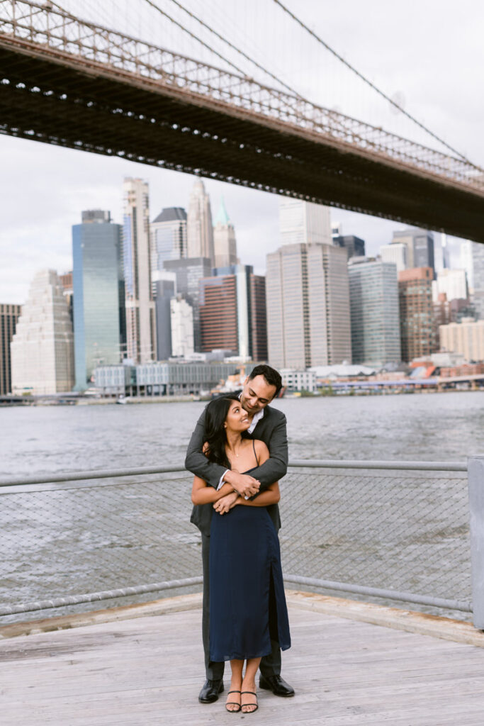 A man with his arms around a woman from behind as they stand on a pier looking out towards the New York City skyline. 
