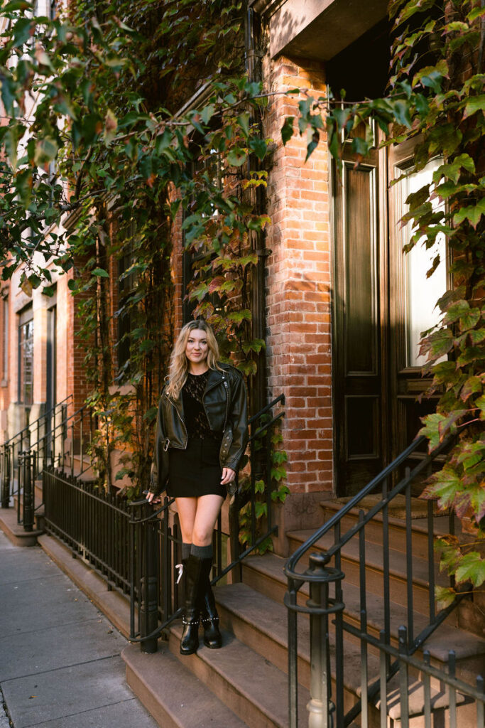 A poised woman in a leather jacket and mini skirt stands on a brownstone's steps, a backdrop of lush ivy enhancing the urban autumn vibe