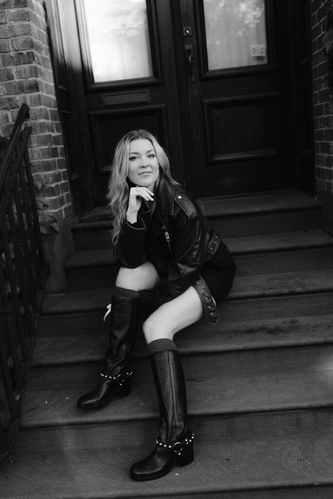 Captured in monochrome, a woman sits on brownstone steps, exuding a timeless elegance in her leather boots and jacket