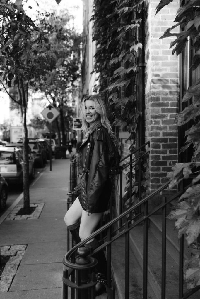 A woman in a black leather jacket and dress smiles over her shoulder on a city street lined with trees and ivy-covered buildings, exuding urban chic