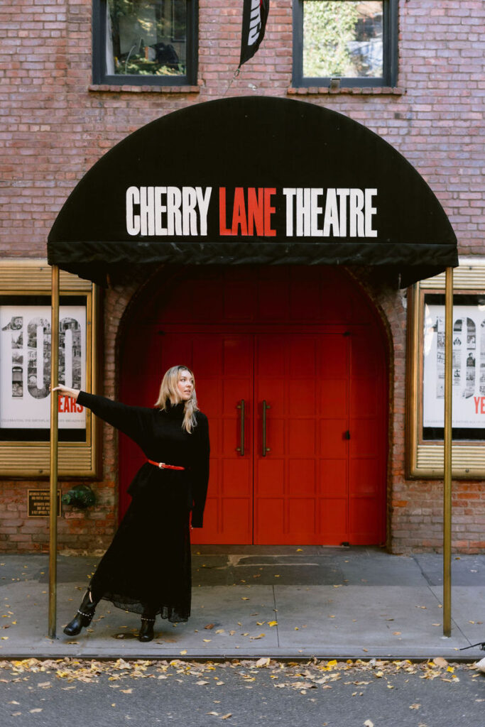 The Cherry Lane Theatre entrance with its iconic red doors is highlighted by a woman in a black dress with a red belt posing under the archway