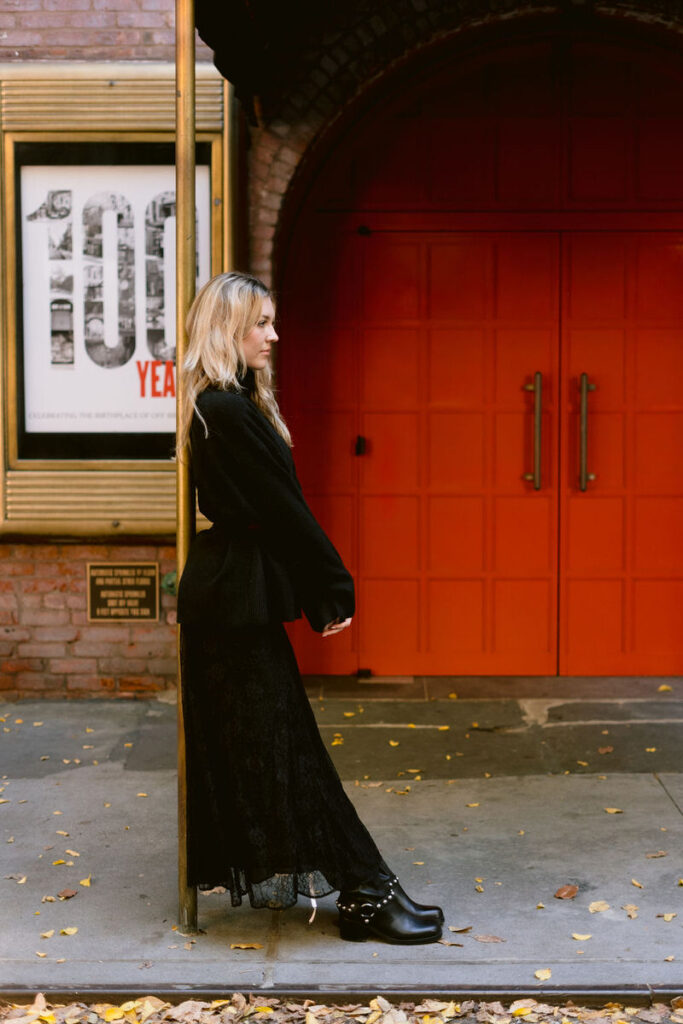 A woman in a chic black ensemble stands thoughtfully by a bright red door, contrasting with the fallen yellow leaves on the sidewalk