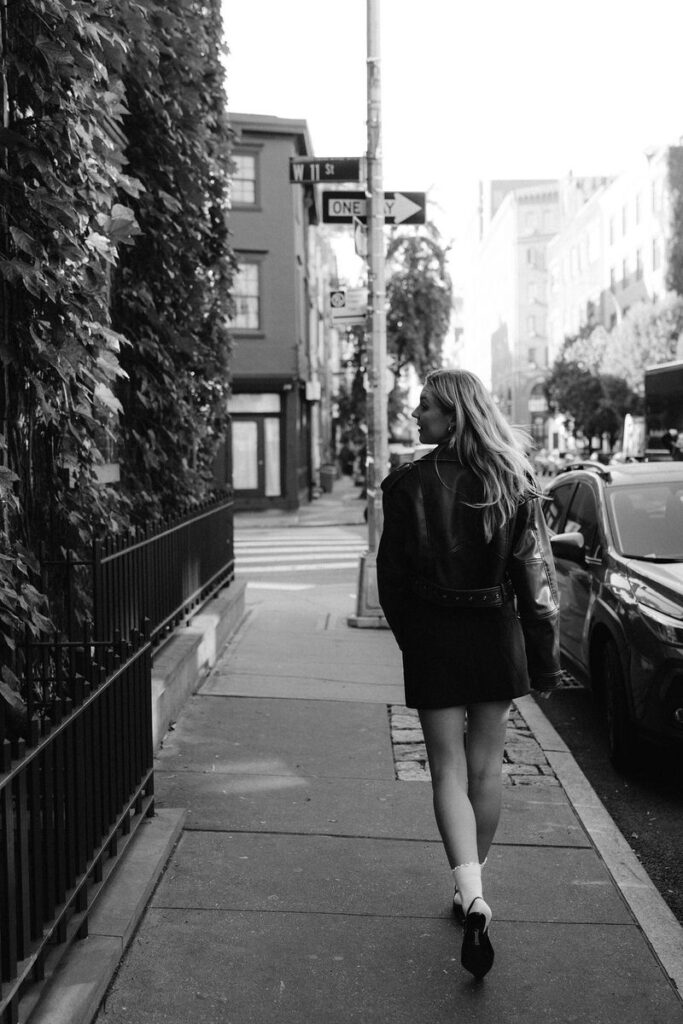 A black and white photo captures a woman in a leather jacket and skirt walking down a sidewalk lined with ivy-covered fences in the West Village, exuding a carefree urban chic vibe