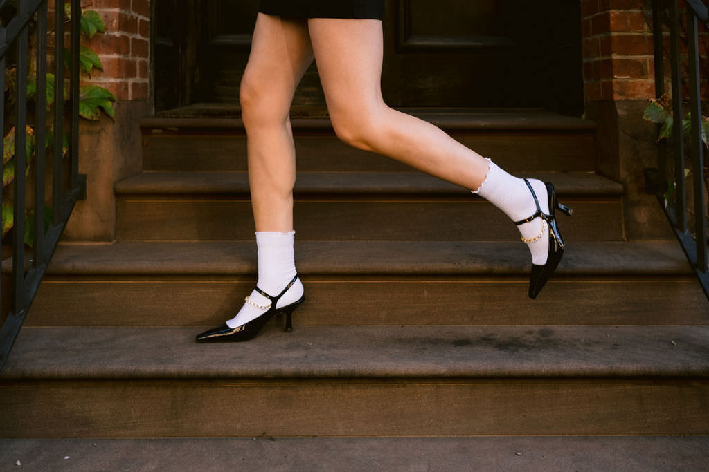 A woman's legs are captured mid-step on the stairs of a brownstone, highlighting her black heels and frilly white socks, embodying a playful yet fashionable spirit