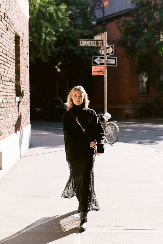 A woman in a black turtleneck and flowing skirt stands confidently on Commerce Street, sunlight highlighting her and the quiet charm of the West Village