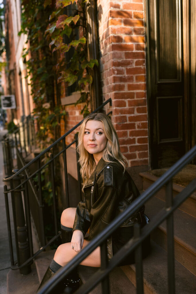 A woman sits contemplatively on the steps of a brownstone, her leather jacket and confident gaze adding to the casual elegance of the West Village ambiance