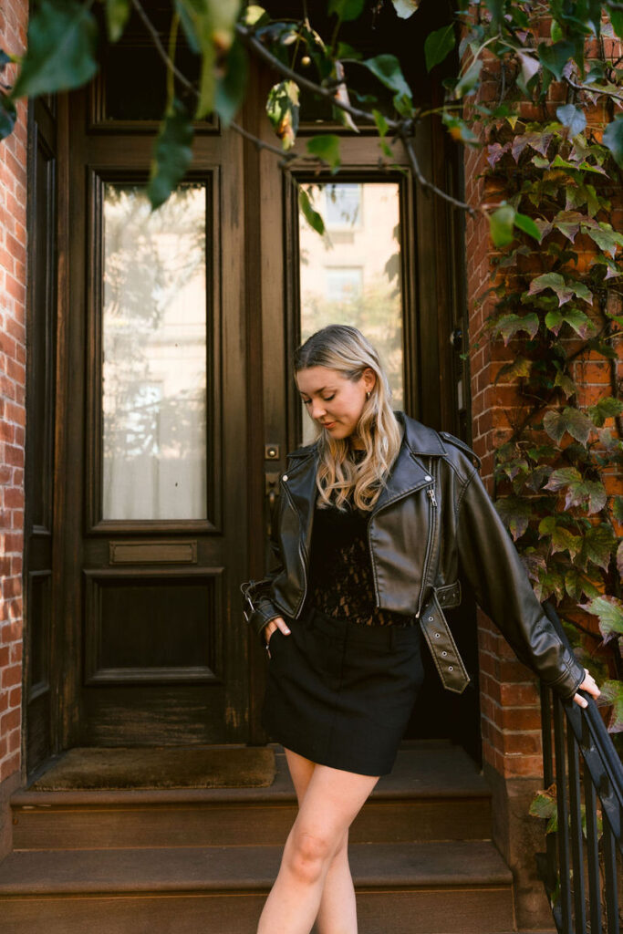 A woman in a stylish black leather jacket and lace top stands on the stoop of a classic brownstone, surrounded by the greenery of ivy leaves in the quiet city morning