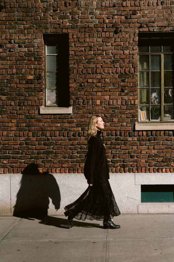A woman in a black outfit walks past a brick wall, her shadow cast beside her in the bright sunlight, reflecting a moment of solitude in the city