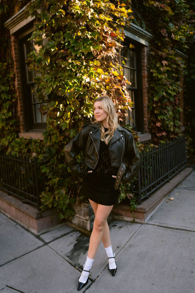 A woman in a leather jacket and skirt stands poised on a New York street corner, with autumn-colored ivy framing the brownstone buildings behind her