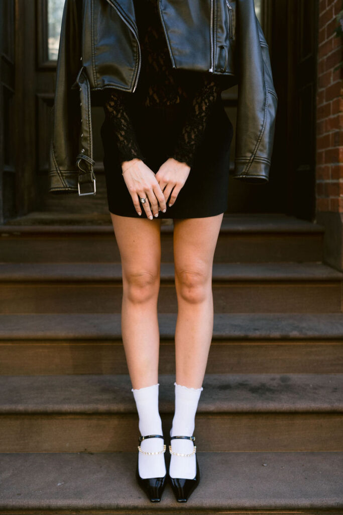 A fashion-focused shot featuring a woman in a lace dress and leather jacket, with a close-up on her hands and chic black heeled shoes with white ruffled socks, on brownstone steps