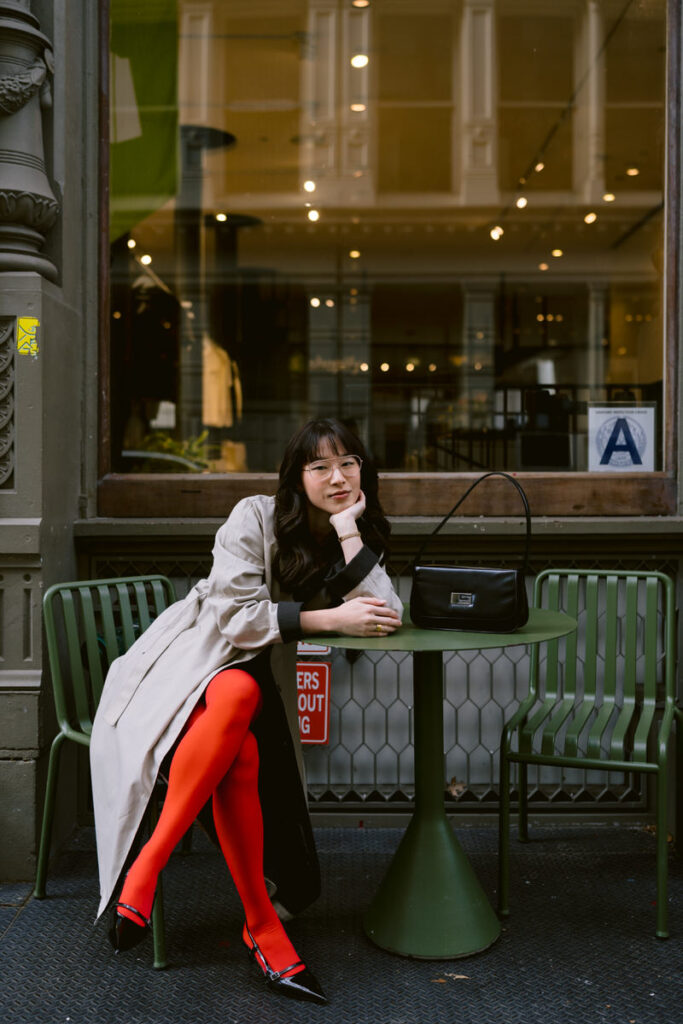 A person seated at an outdoor café table, elegantly resting their chin on their hand, with a black Gucci shoulder bag placed on the table and bright red tights paired with black heels